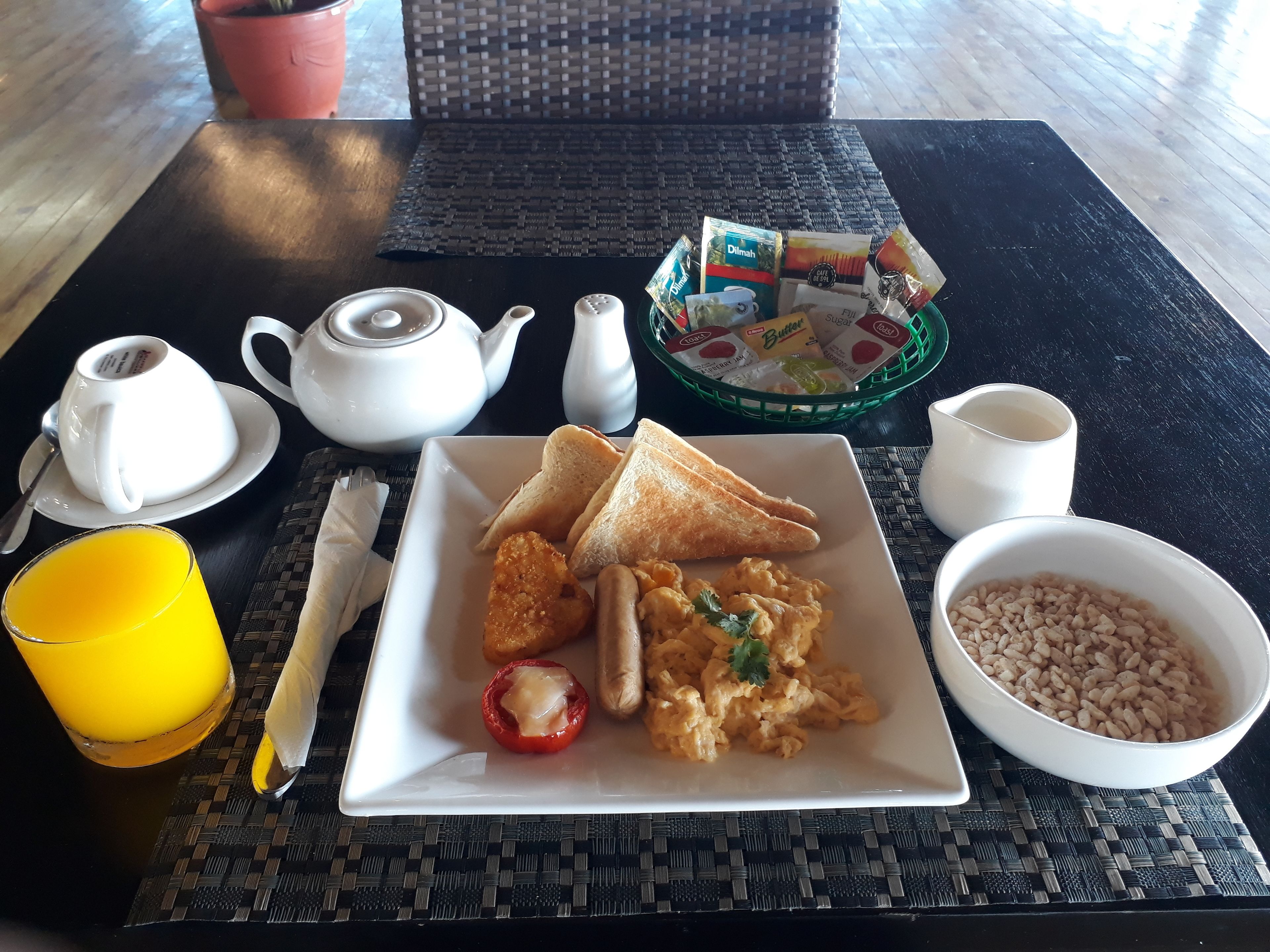 A breakfast setting with toast, scrambled eggs, a tomato, beans, a teapot, cups, and a juice glass on a table.