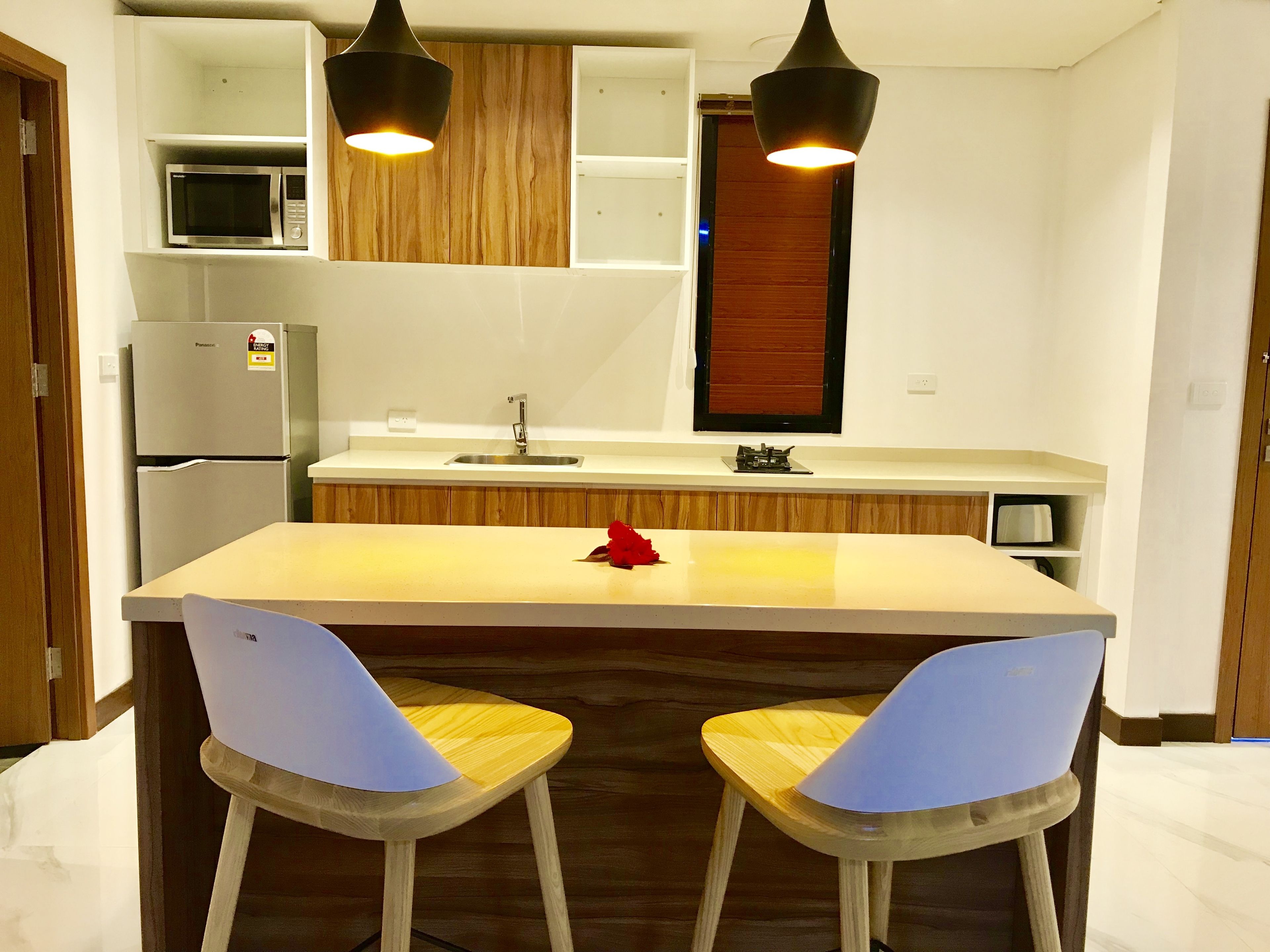 A modern kitchen with a central island, two bar stools, a microwave, a refrigerator, and pendant lighting.