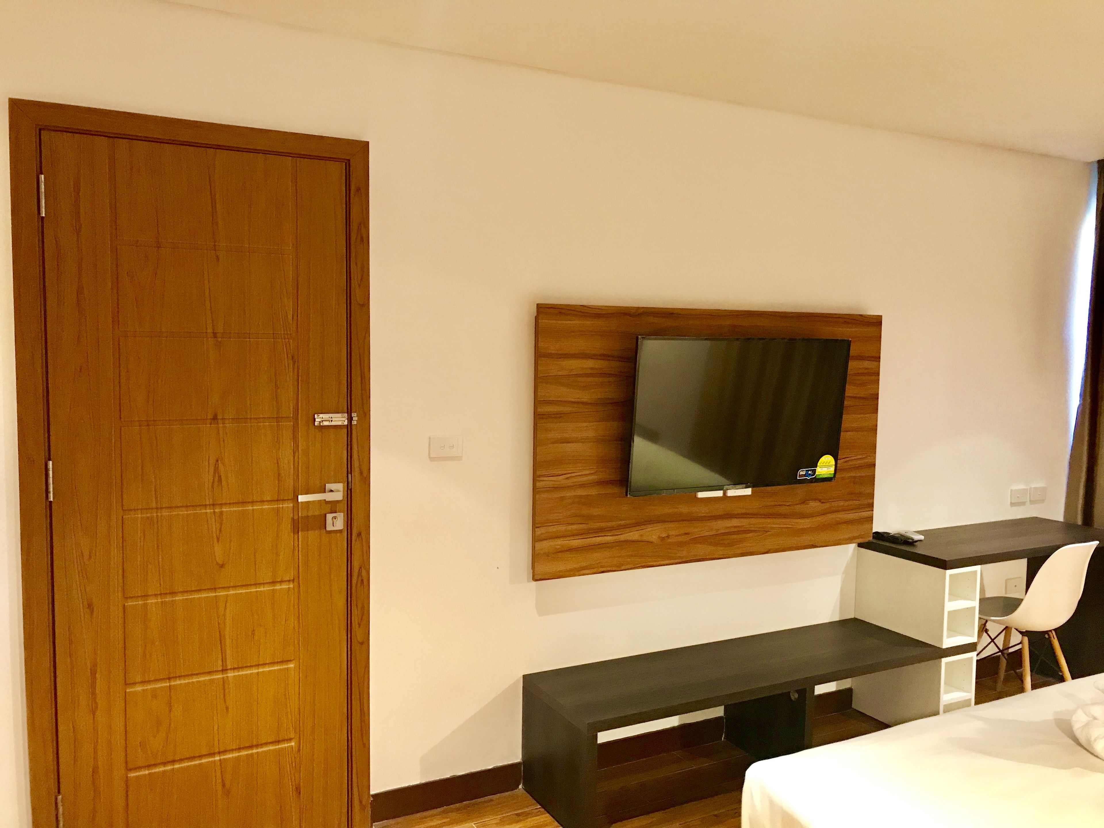 Une chambre d’hôtel avec une porte en bois, un téléviseur à écran plat fixé sur un panneau en bois, un bureau avec une chaise et un lit avec du linge de lit blanc.