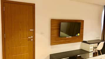 Une chambre d’hôtel avec une porte en bois, un téléviseur à écran plat fixé sur un panneau en bois, un bureau avec une chaise et un lit avec du linge de lit blanc.