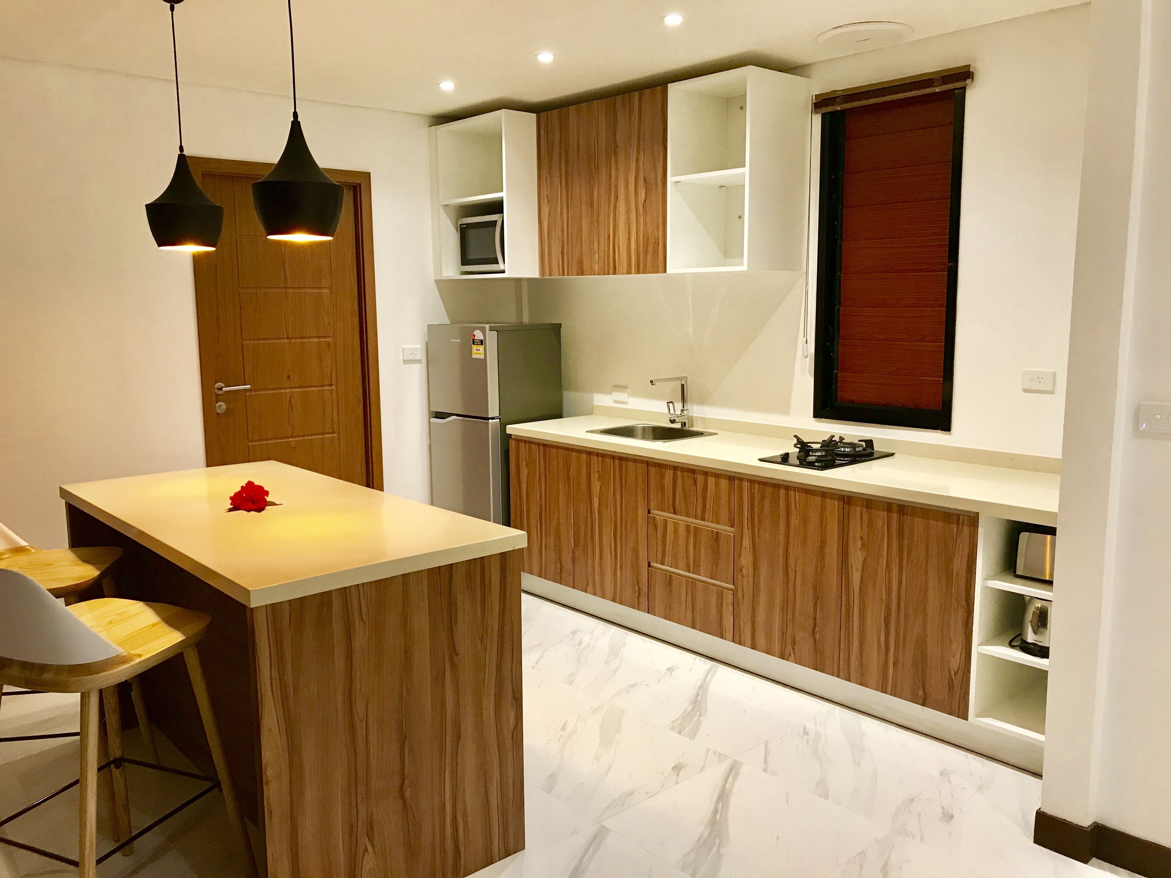 A compact kitchen with wooden cabinets, a stainless steel refrigerator, a sink, and a small dining area with a chair.