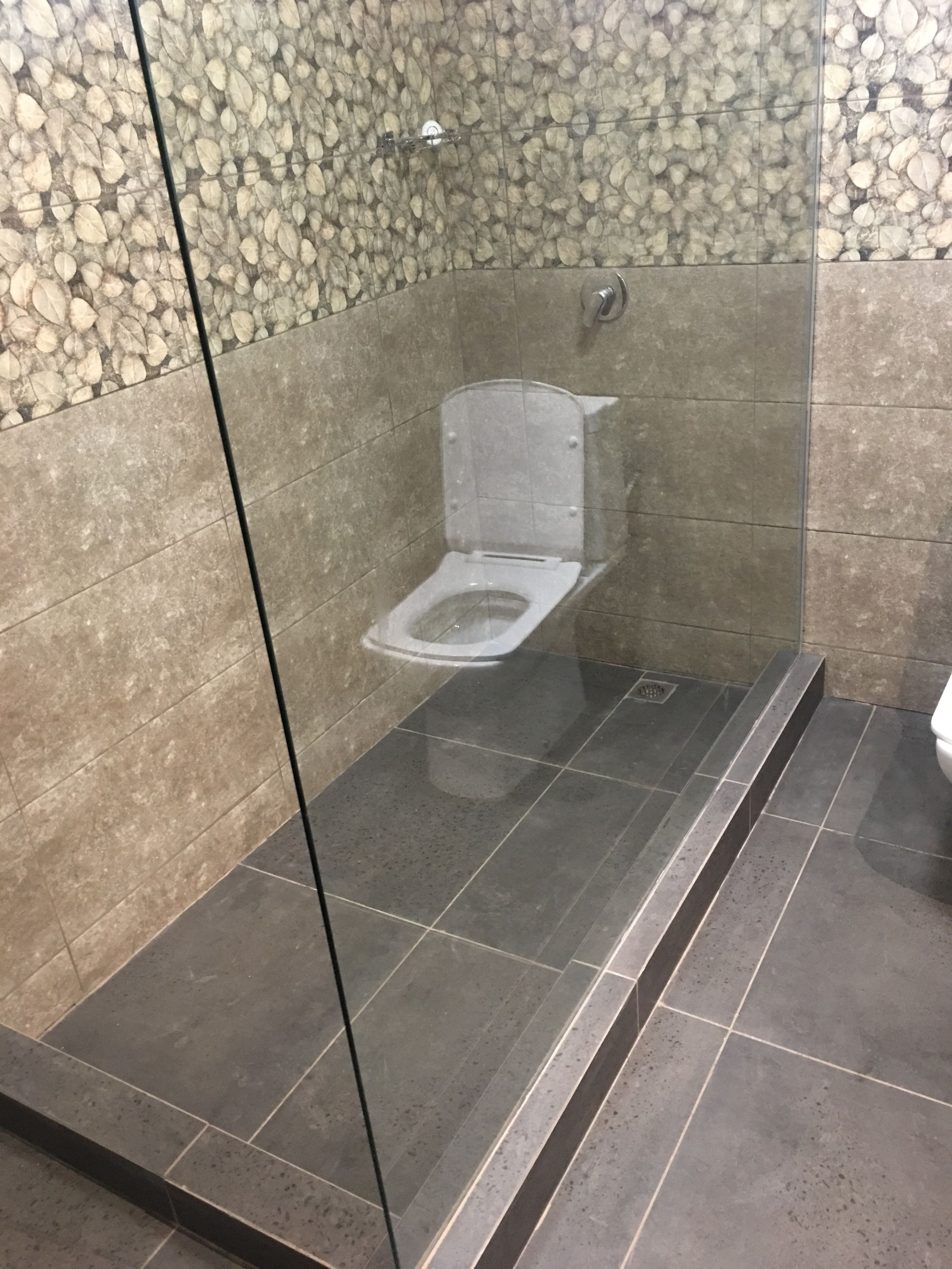 Un baño moderno con ducha envuelta en vidrio, paredes con diseño de piedra y piso de baldosas oscuras.