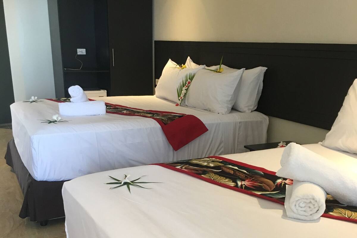 Habitación de hotel con dos camas, cada una con ropa blanca y almohadas decorativas. Se observa un armario y una pequeña mesa en la habitación.