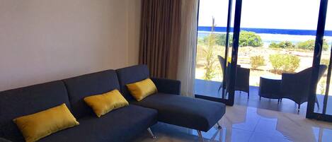 Een moderne woonkamer met een donkerblauwe bank en gele kussens, een glazen deur die leidt naar een terras met uitzicht op het strand.