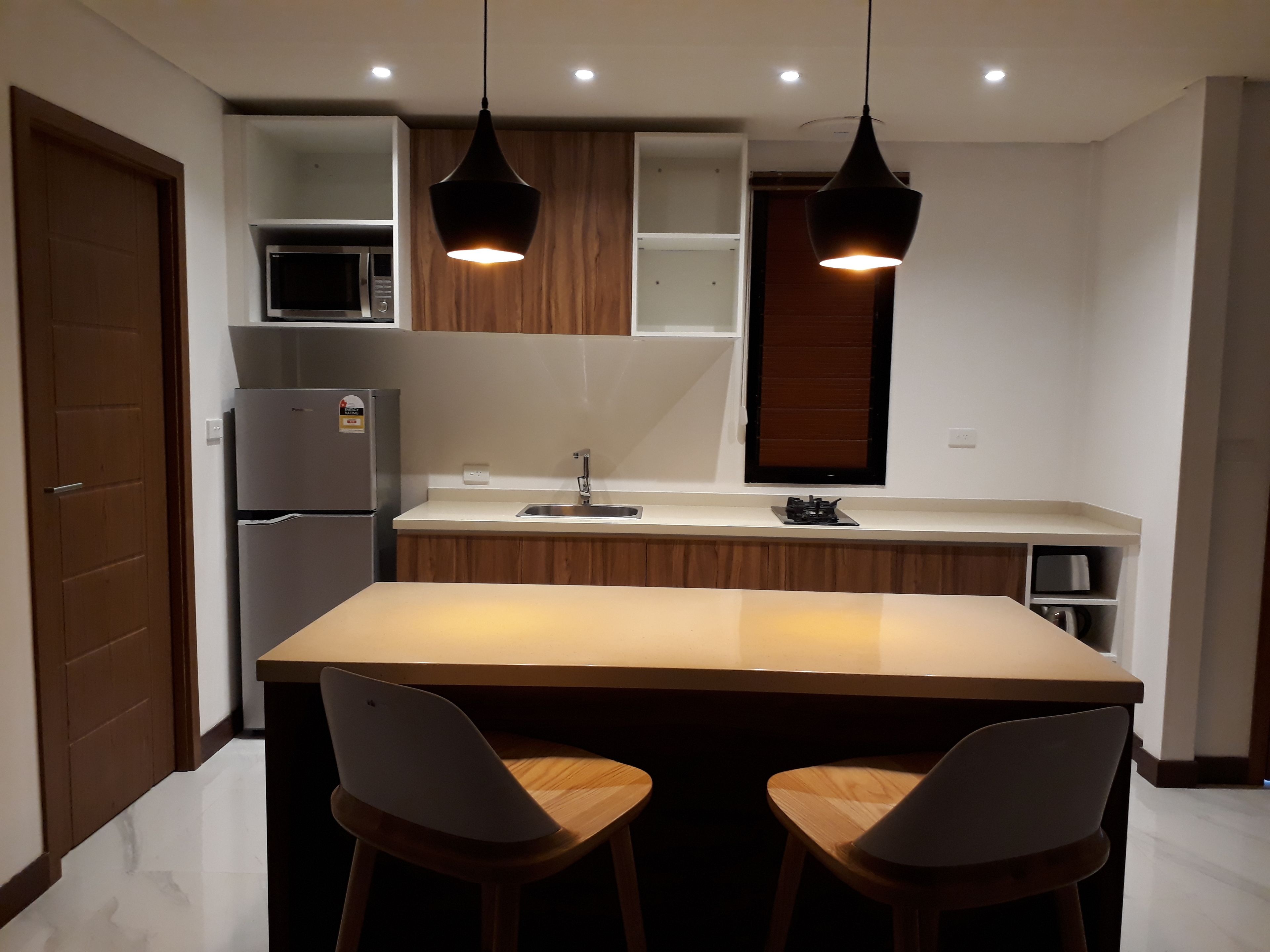 A modern kitchen with a central island, two bar stools, wooden cabinets, a built-in microwave, and pendant lighting.