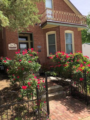 A brick building with a balcony, a sign indicating it's a guest house, and blooming red flowers in the front yard.