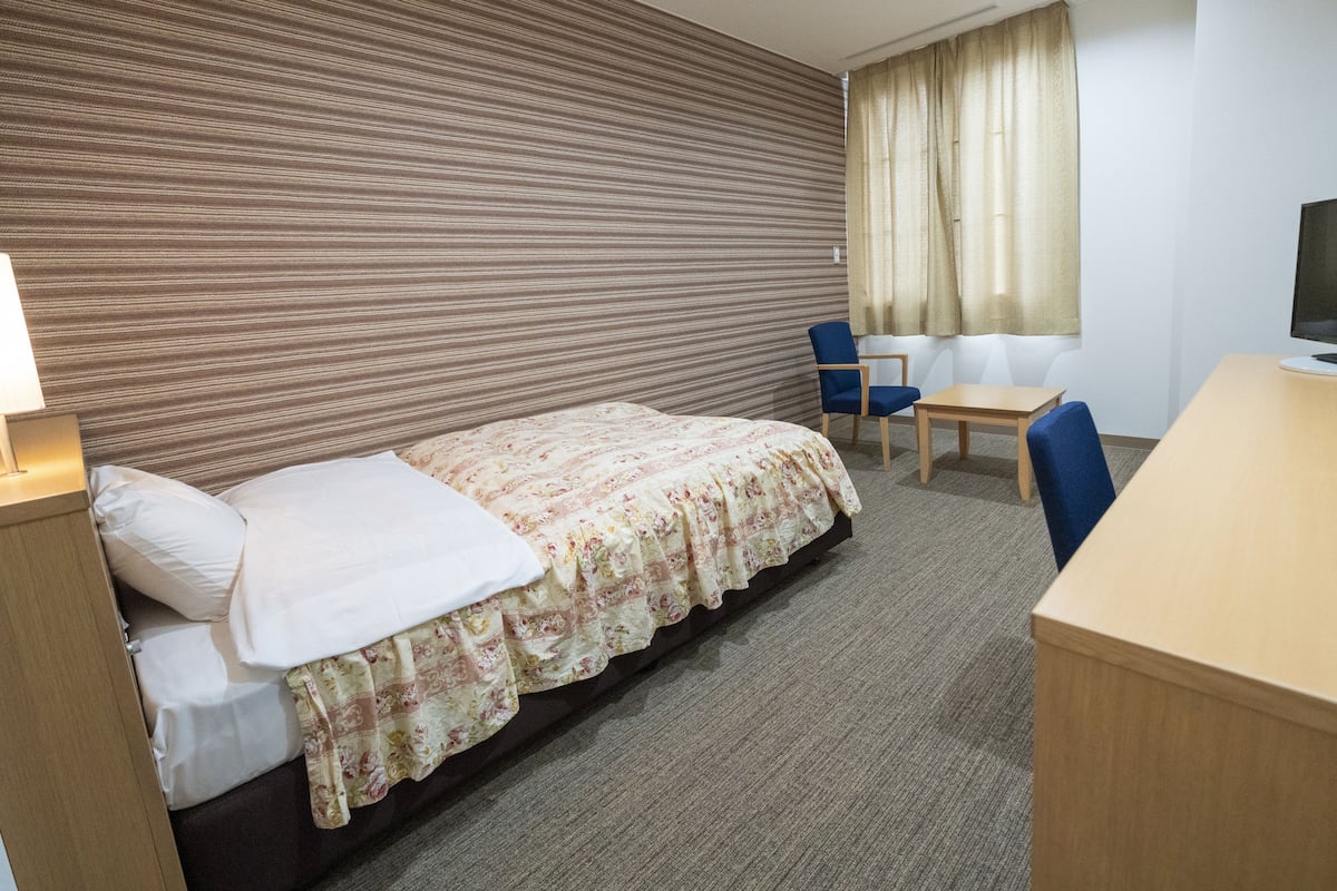 Une chambre d’hôtel avec un lit, un bureau, une chaise, une télévision et une fenêtre avec des rideaux.