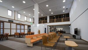 Um hall amplo com teto alto, várias áreas de estar incluindo um sofá laranja comprido e cadeiras de madeira, e um balcão no segundo andar com guarda-corpo de madeira.