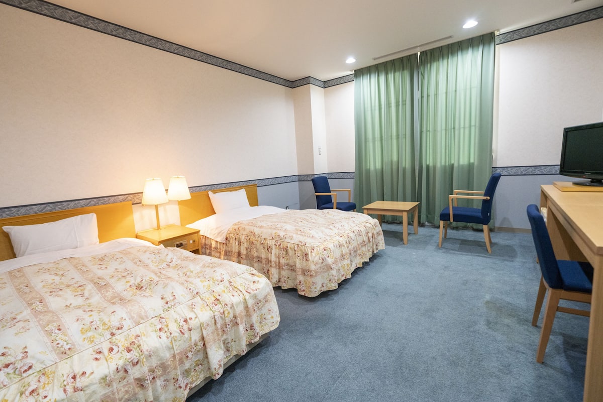 Une chambre d’hôtel avec deux lits, un bureau, une chaise, une télévision et une fenêtre avec des rideaux.