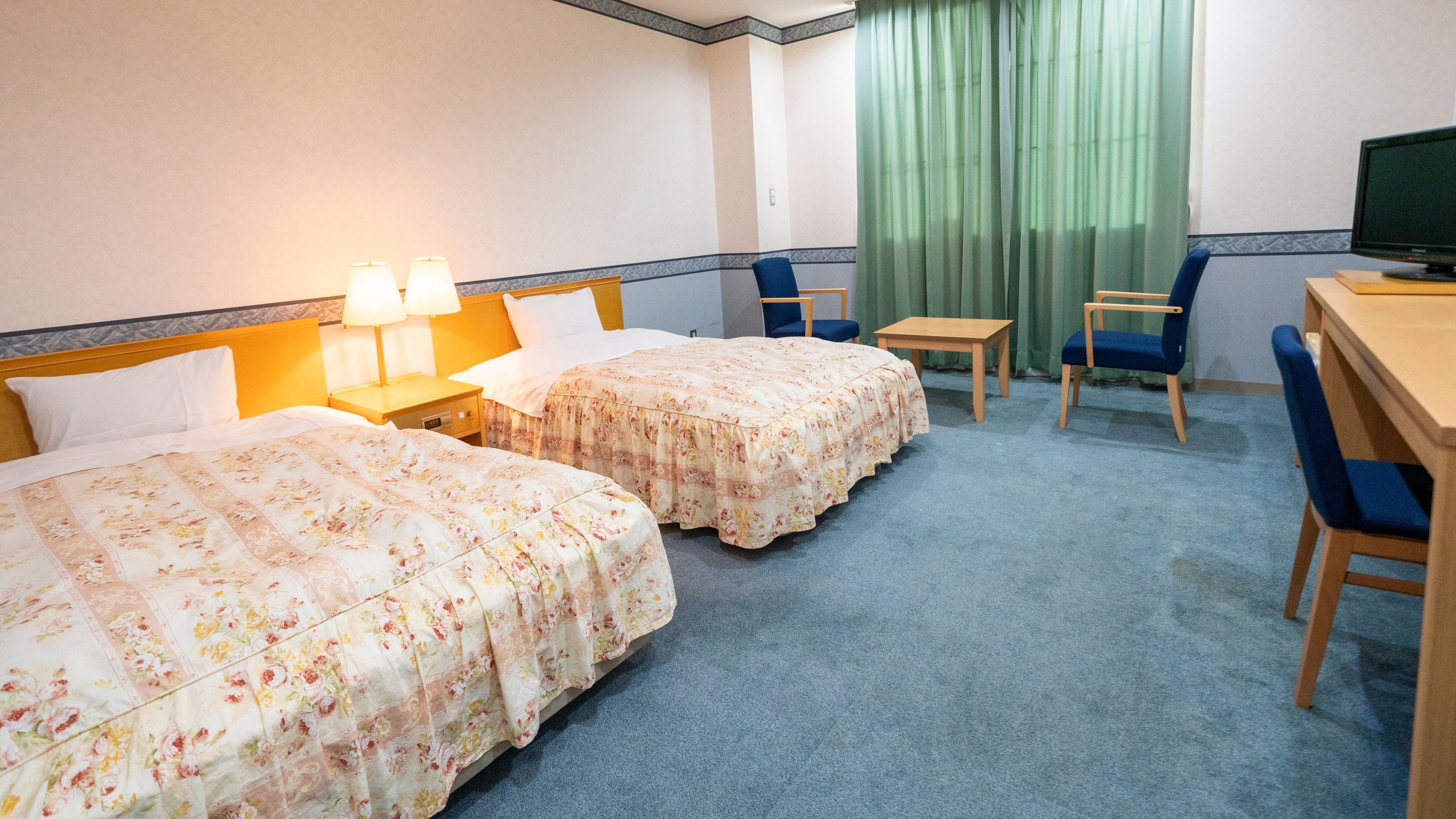 Ein Hotelzimmer mit zwei Betten, einem Schreibtisch, einem Stuhl, einem Fernseher und einem Fenster mit Vorhängen.
