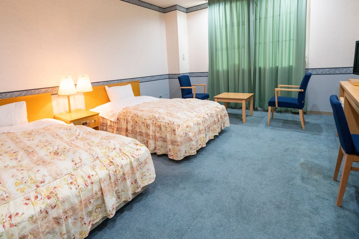 Une chambre d’hôtel avec deux lits, un bureau, une chaise, une télévision et une fenêtre avec des rideaux.