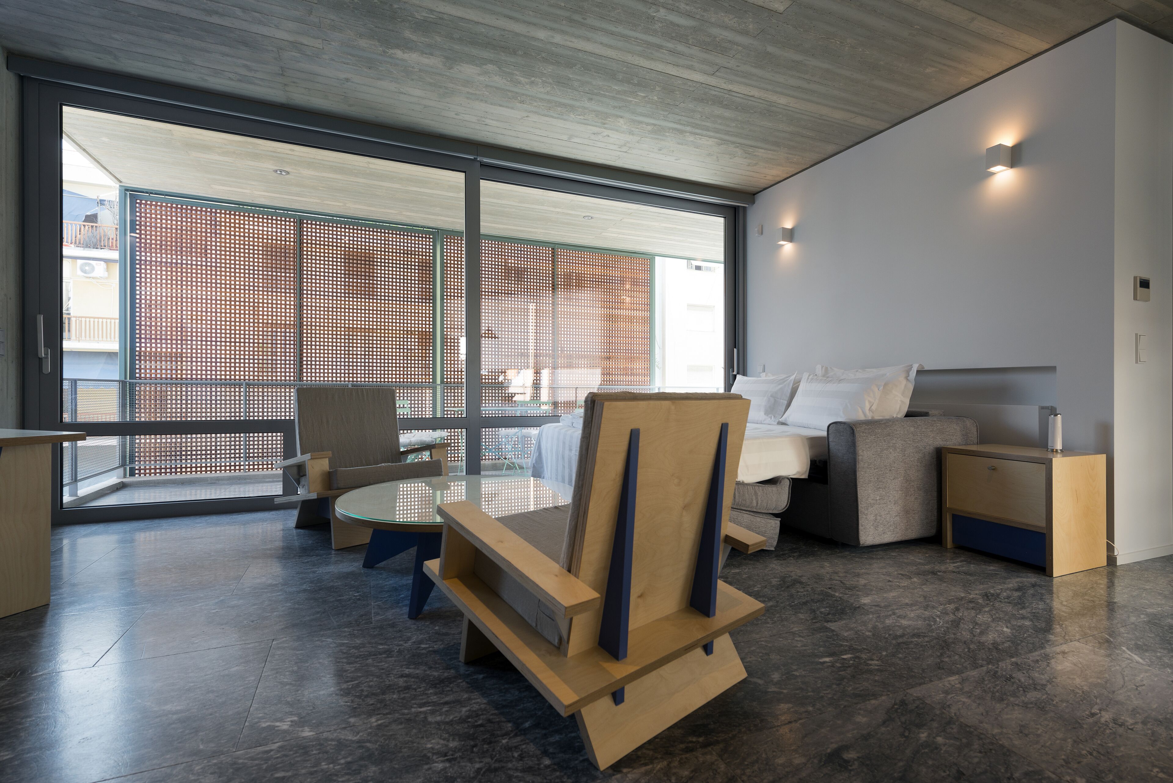 A modern living room with a sofa, a wooden chair, a small table, and a balcony with a view of a building.