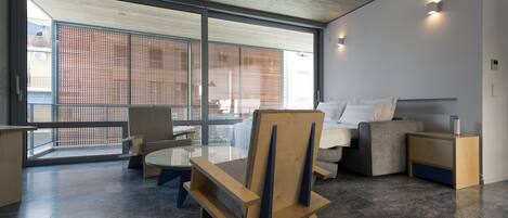 A modern living room with a sofa, a wooden chair, a small table, and a balcony with a view of a building.