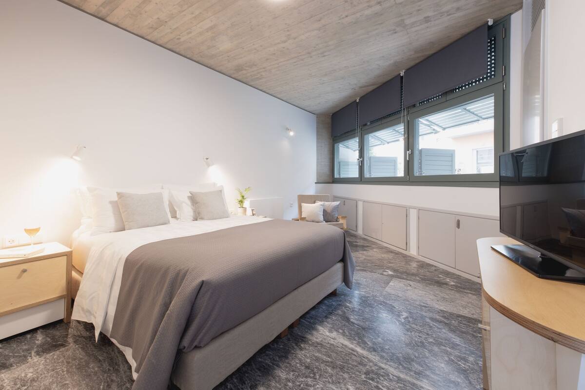 A modern bedroom with a large bed, bedside tables, a TV, and a window with blinds.