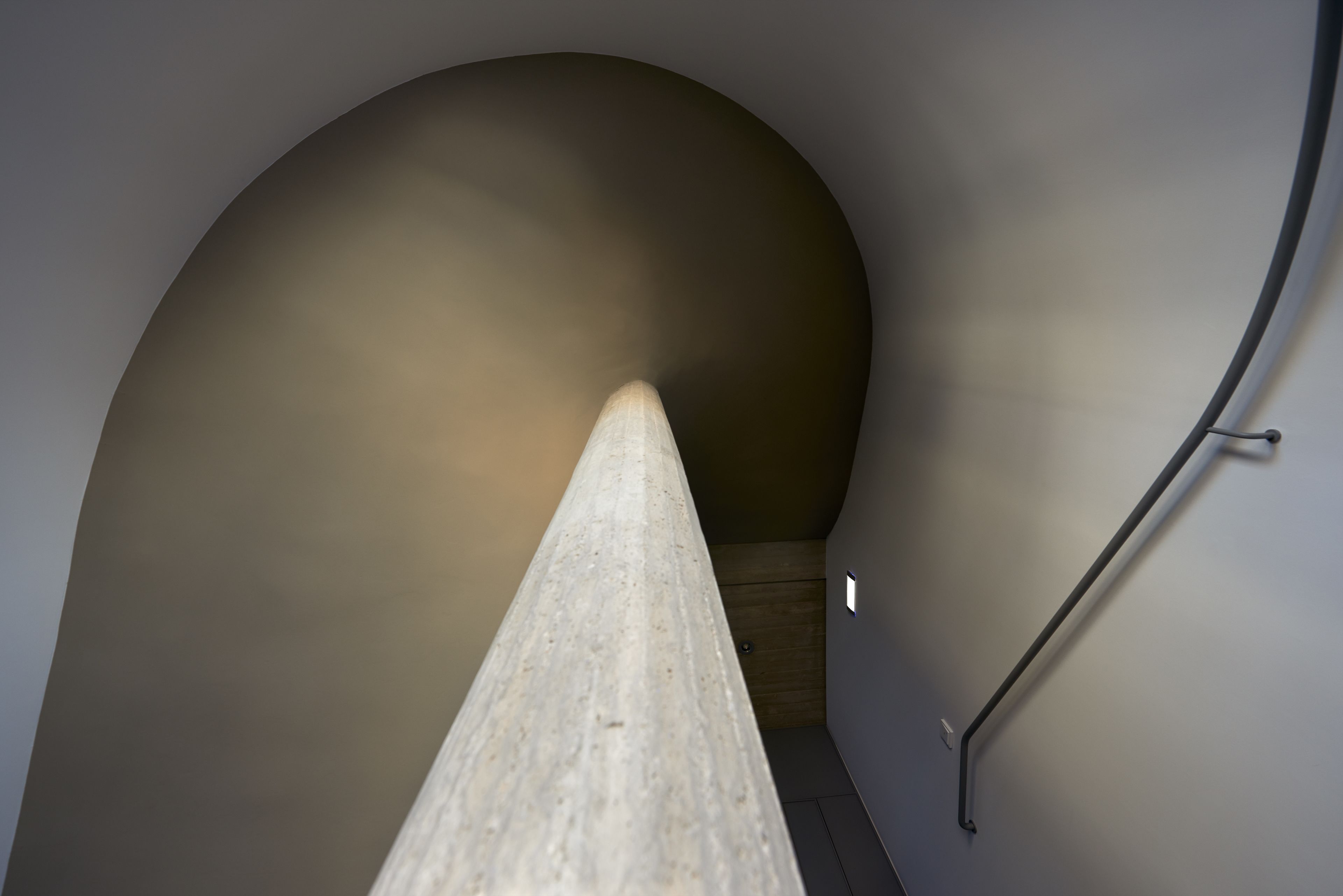 A narrow, curved stairway with a wooden handrail and a light fixture on the wall.