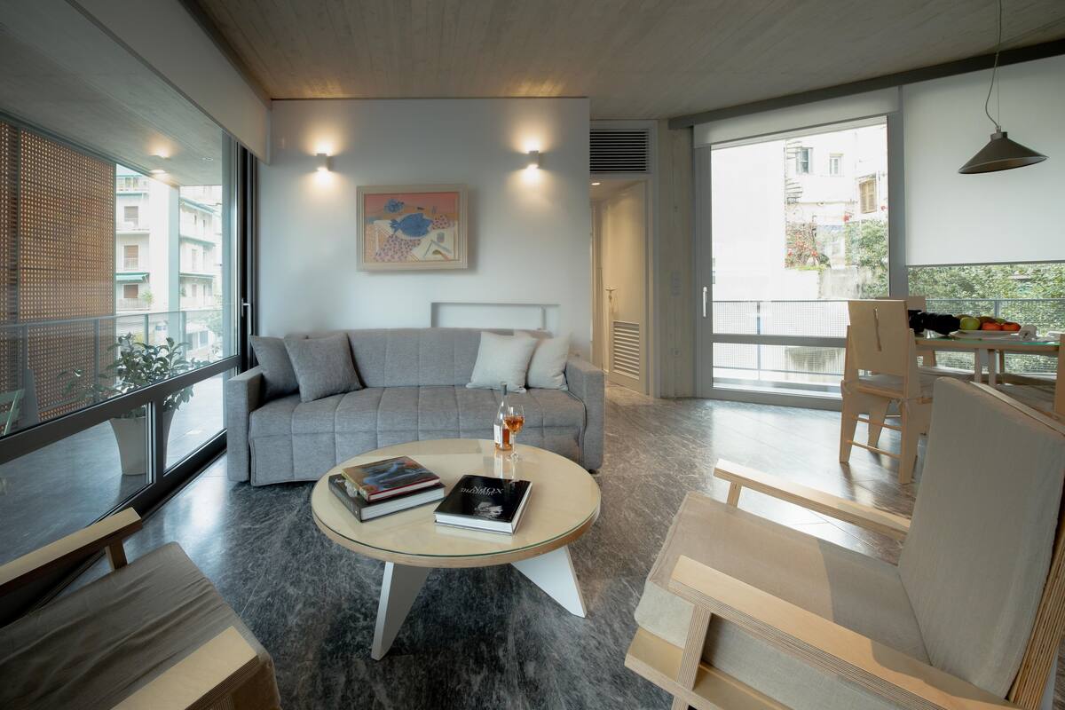 A modern living room with a grey sofa, a round coffee table with books, and a large window offering a view of buildings.