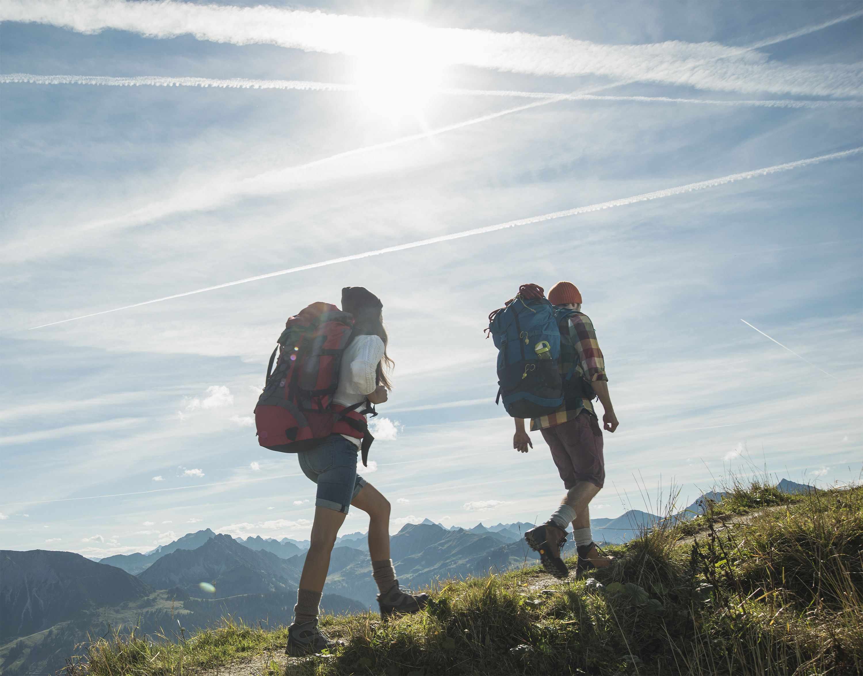Two hikers with backpacks on a mountain trail with a clear sky and distant mountains.