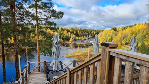 A wooden deck with a railing, overlooking a river and autumn-colored trees. - Björnforsens Turist & Konferens Hotell (Bjorna)