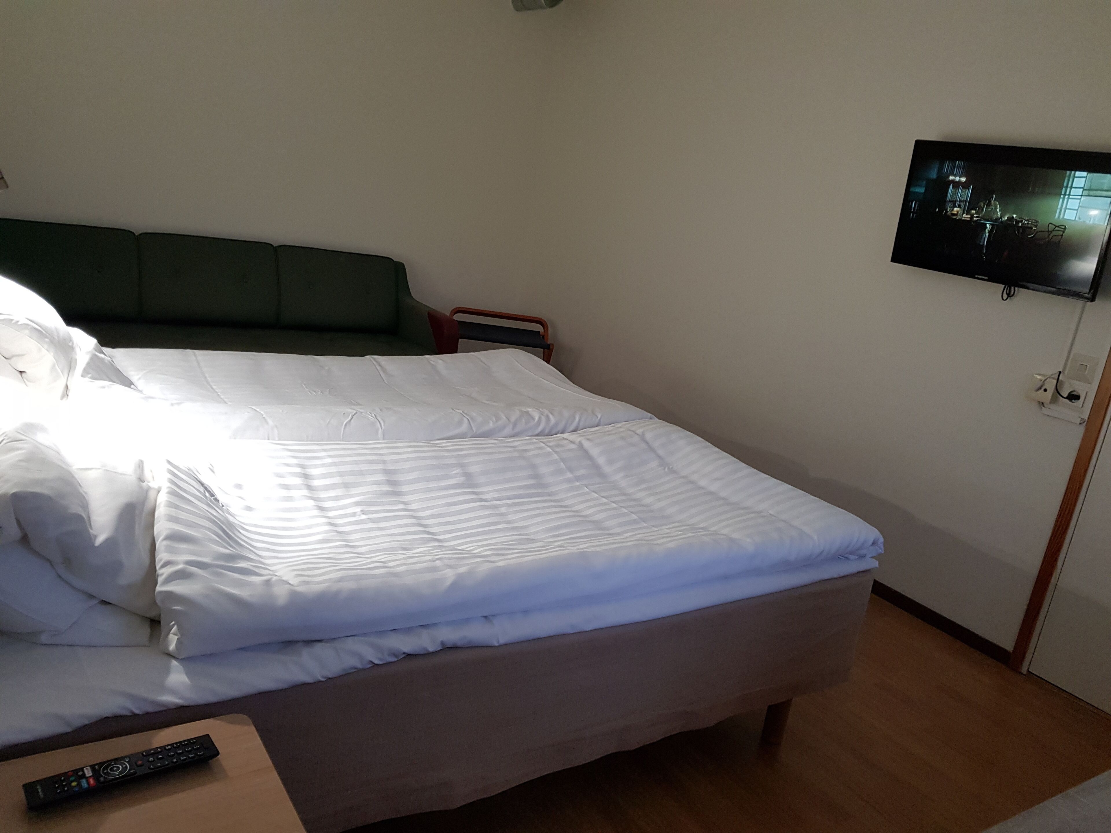 Siististi pedattu sänky valkoisilla lakanoilla, kaukosäädin puisen yöpöydän päällä ja seinälle asennettu televisio.