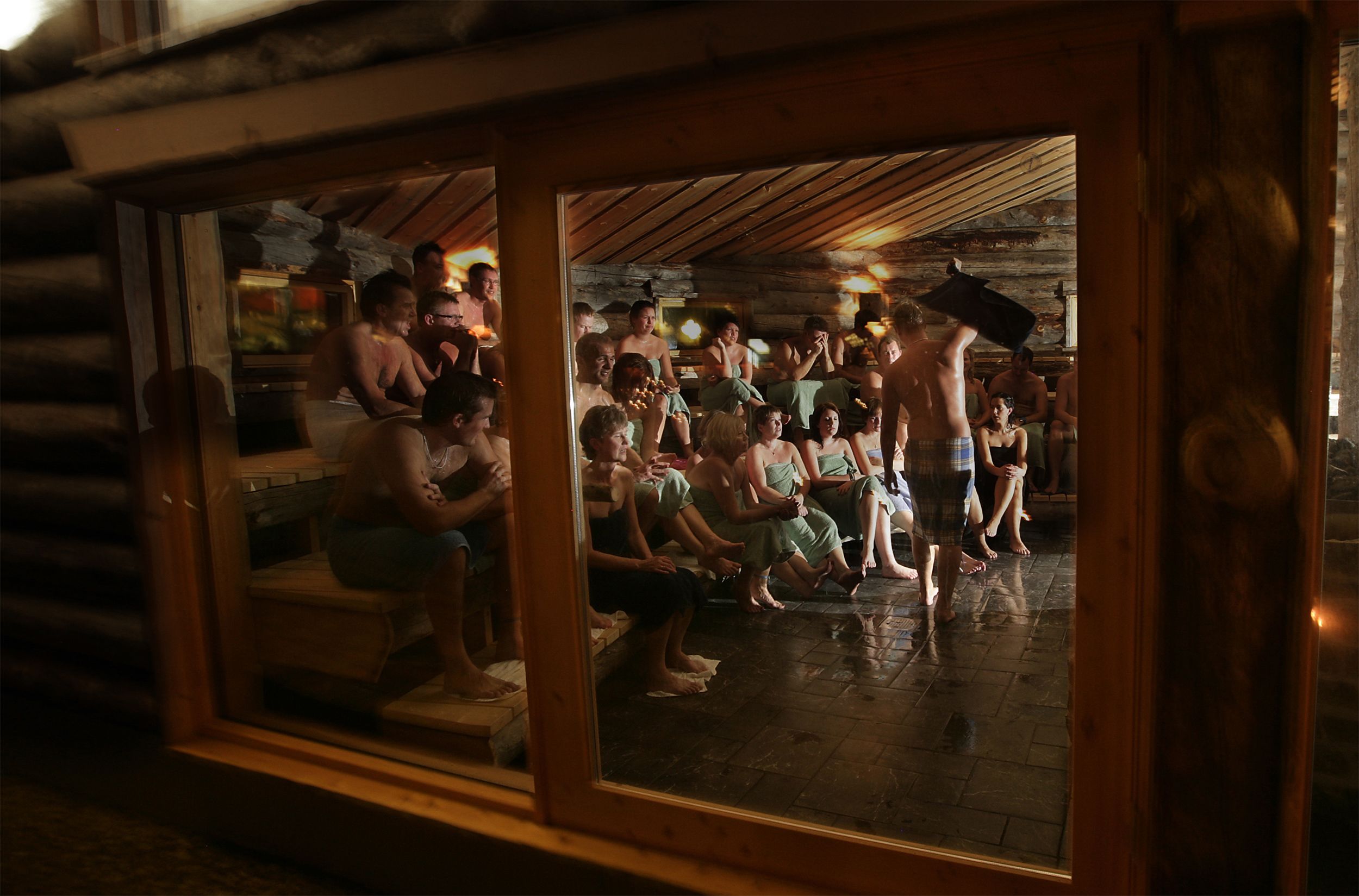 Interior de cabana de tronco com um grupo de pessoas reunidas no centro, algumas em pé e outras sentadas.