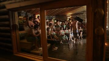 Interior de cabana de tronco com um grupo de pessoas reunidas no centro, algumas em pé e outras sentadas.