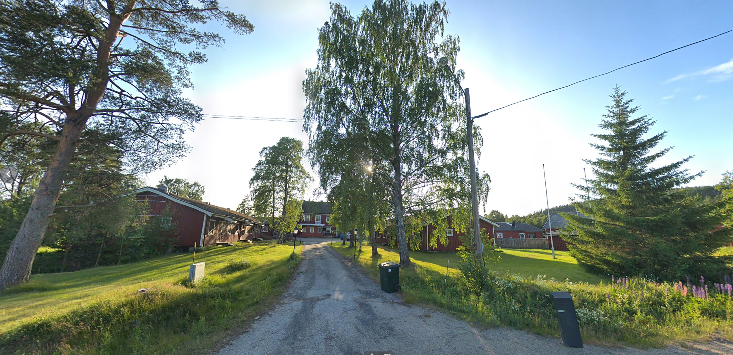 A gravel road leading to a row of red buildings, surrounded by trees and greenery.