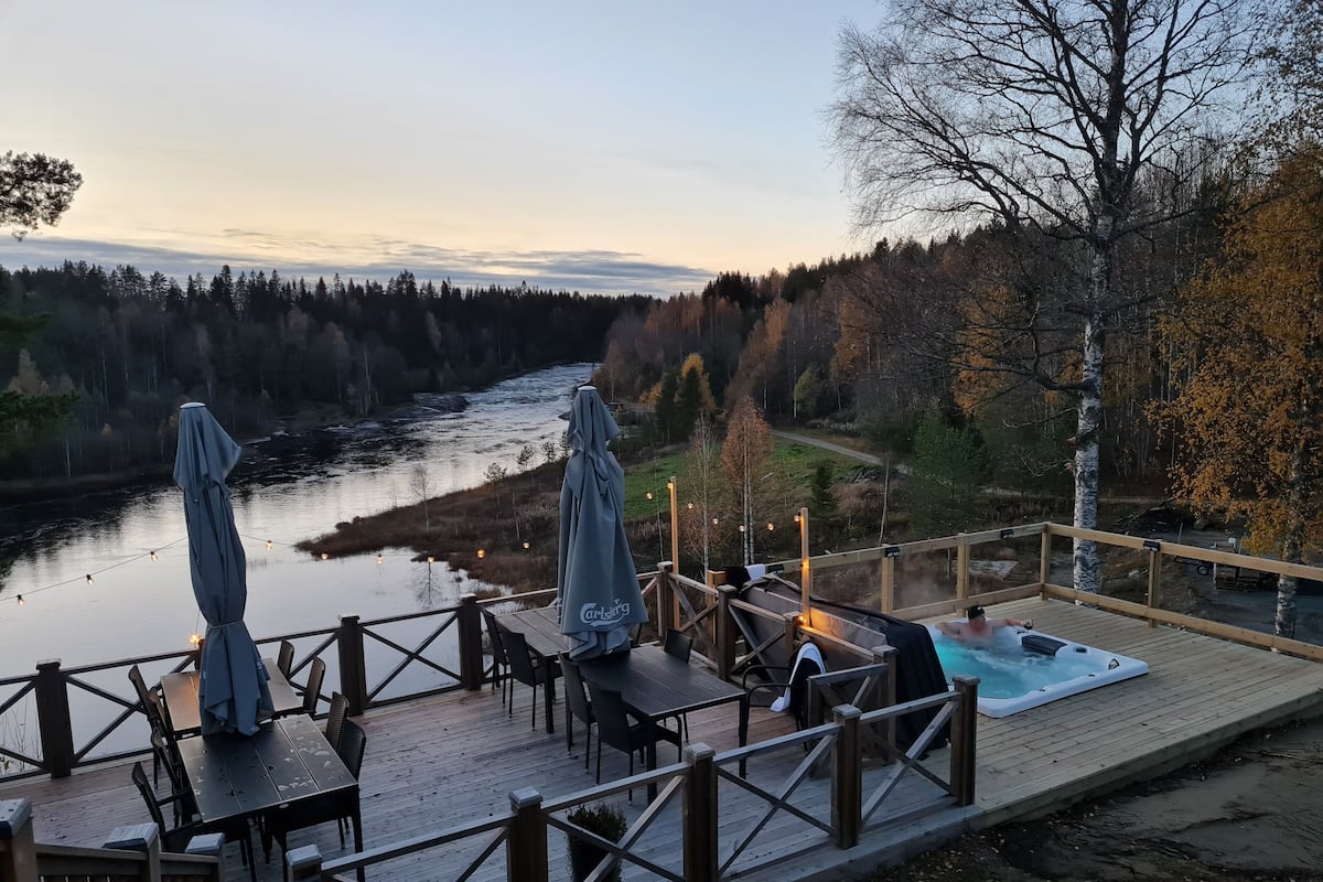 Une terrasse en bois avec des sièges d’extérieur et un jacuzzi, le tout offrant une vue sur une rivière et une forêt.