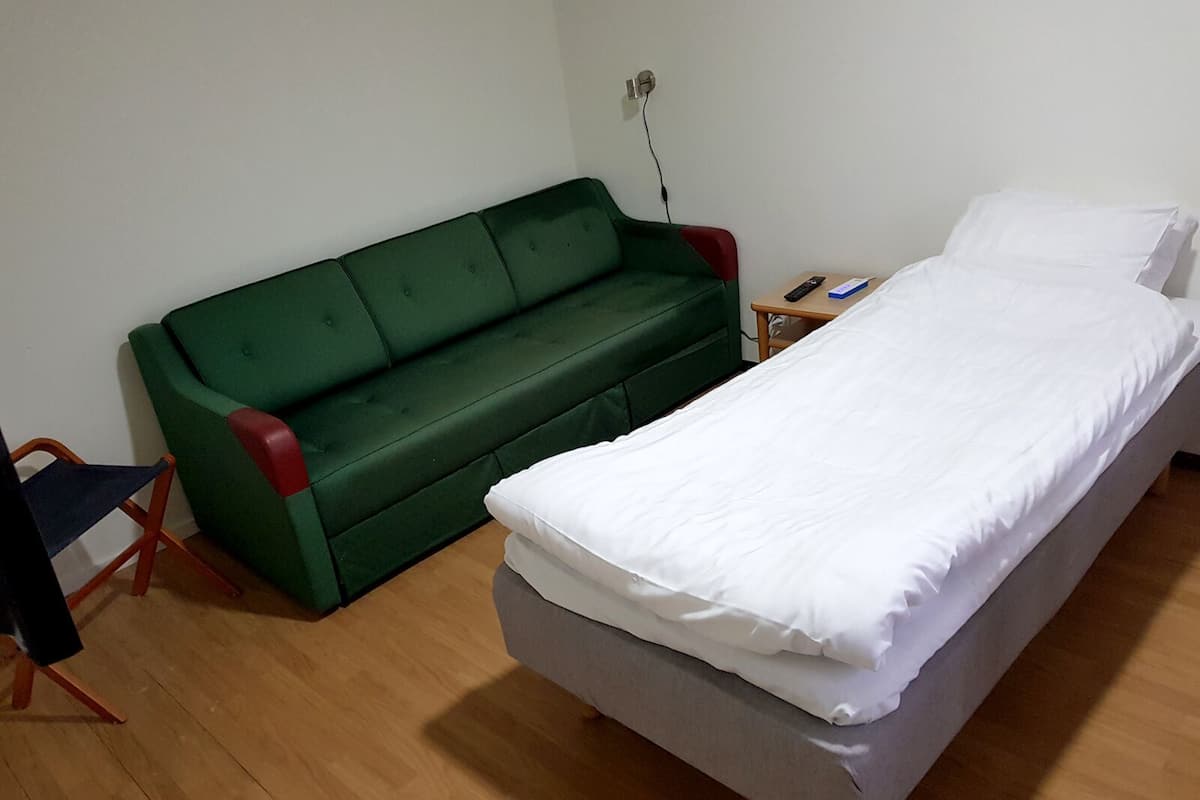 Une petite pièce simple, avec un lit simple, un canapé vert et une petite table sur laquelle se trouve une télécommande.