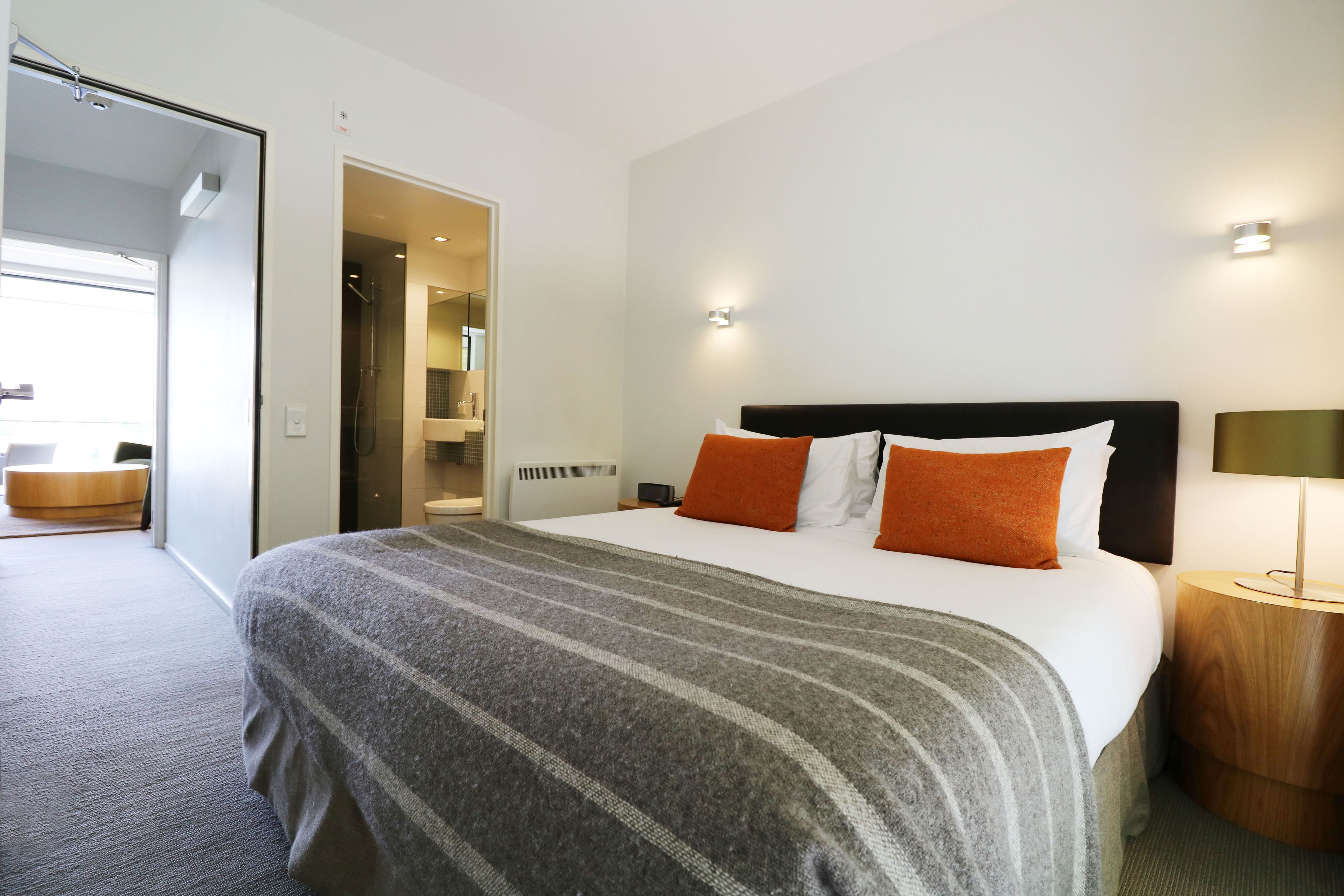 Quarto de hotel com uma cama grande, uma mesa de cabeceira de madeira e um banheiro visível por uma porta aberta.