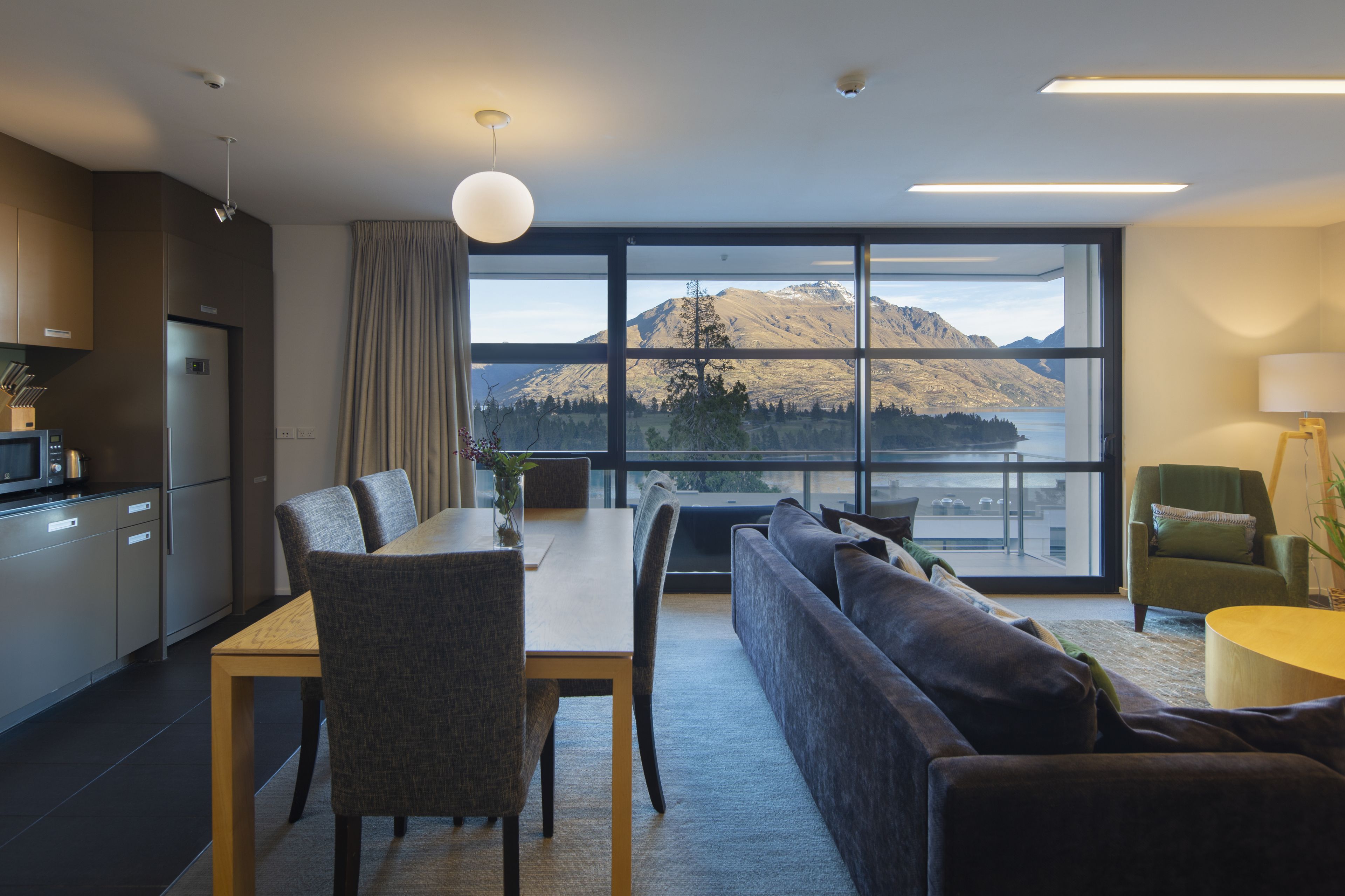 A modern kitchen with a dining area, a sofa, and a view of mountains through large windows.