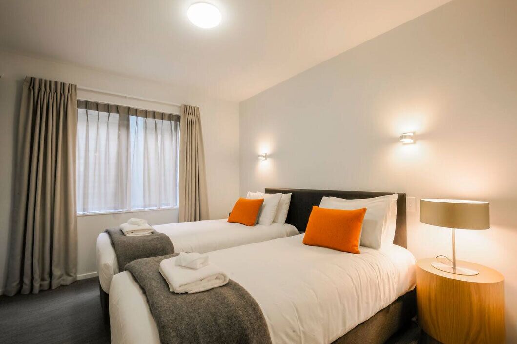 Une chambre d’hôtel avec deux lits, une table de chevet, une lampe et une fenêtre avec des rideaux.