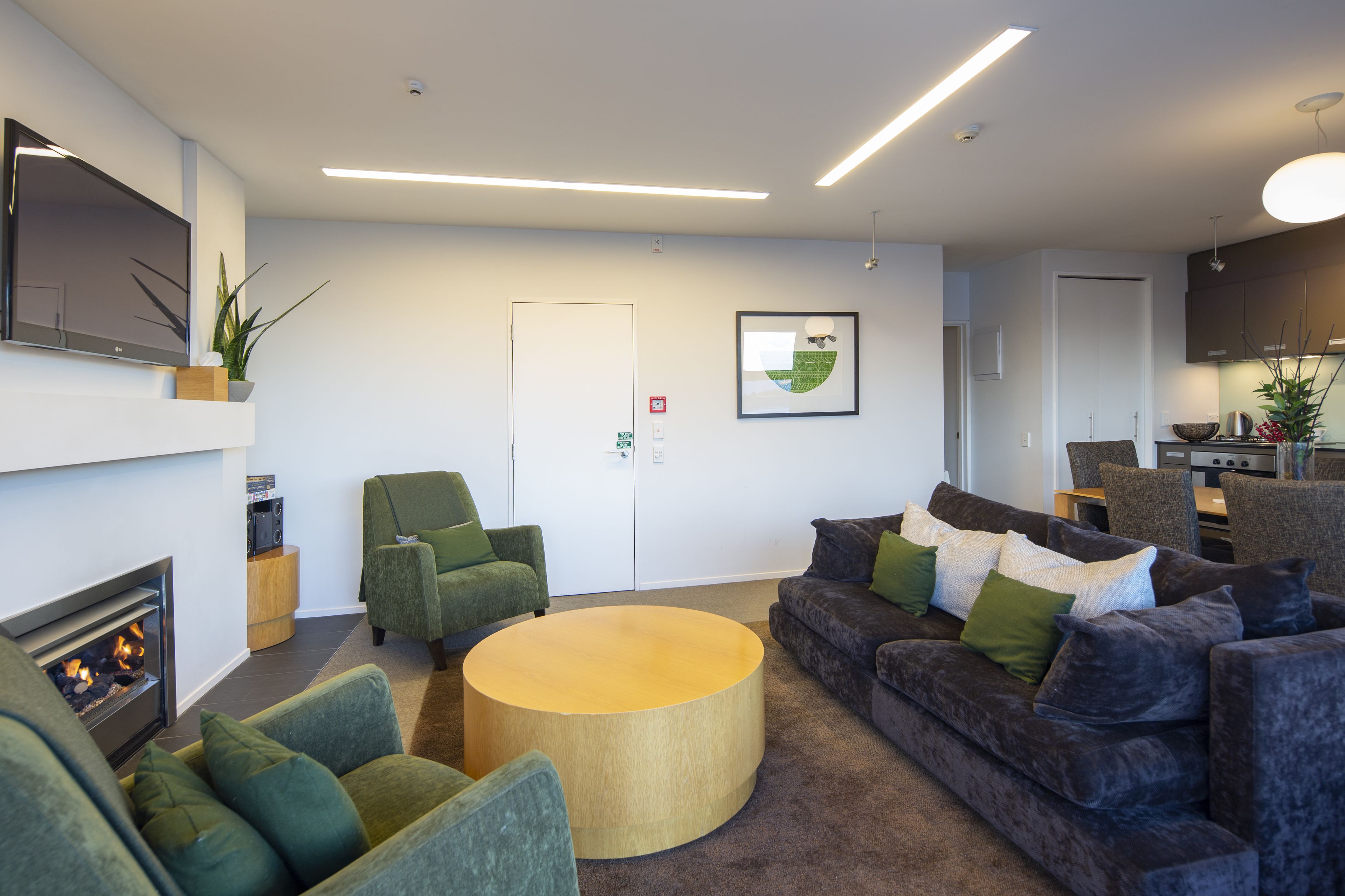 A modern living room with a fireplace, comfortable seating, a flat-screen TV, and a kitchen area in the background.