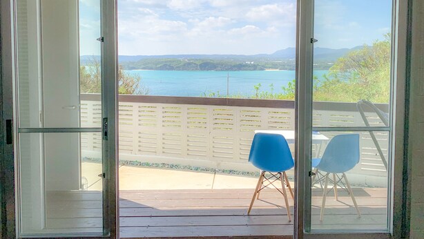 A room with a sliding glass door leading to an outdoor deck with two blue chairs and a view of a lake and mountains. - Resort Villa Kouri island Aqua Blue (Nakijin)