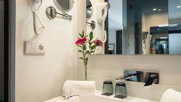 A modern bathroom with a white sink, a mirror, a hairdryer, and a vase with flowers.