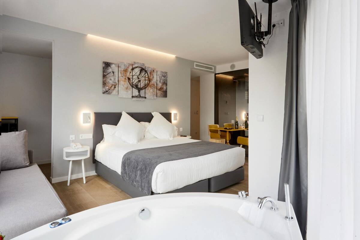 A modern hotel room with a large bed, a separate sitting area, a bathroom with a bathtub, and a dining area.
