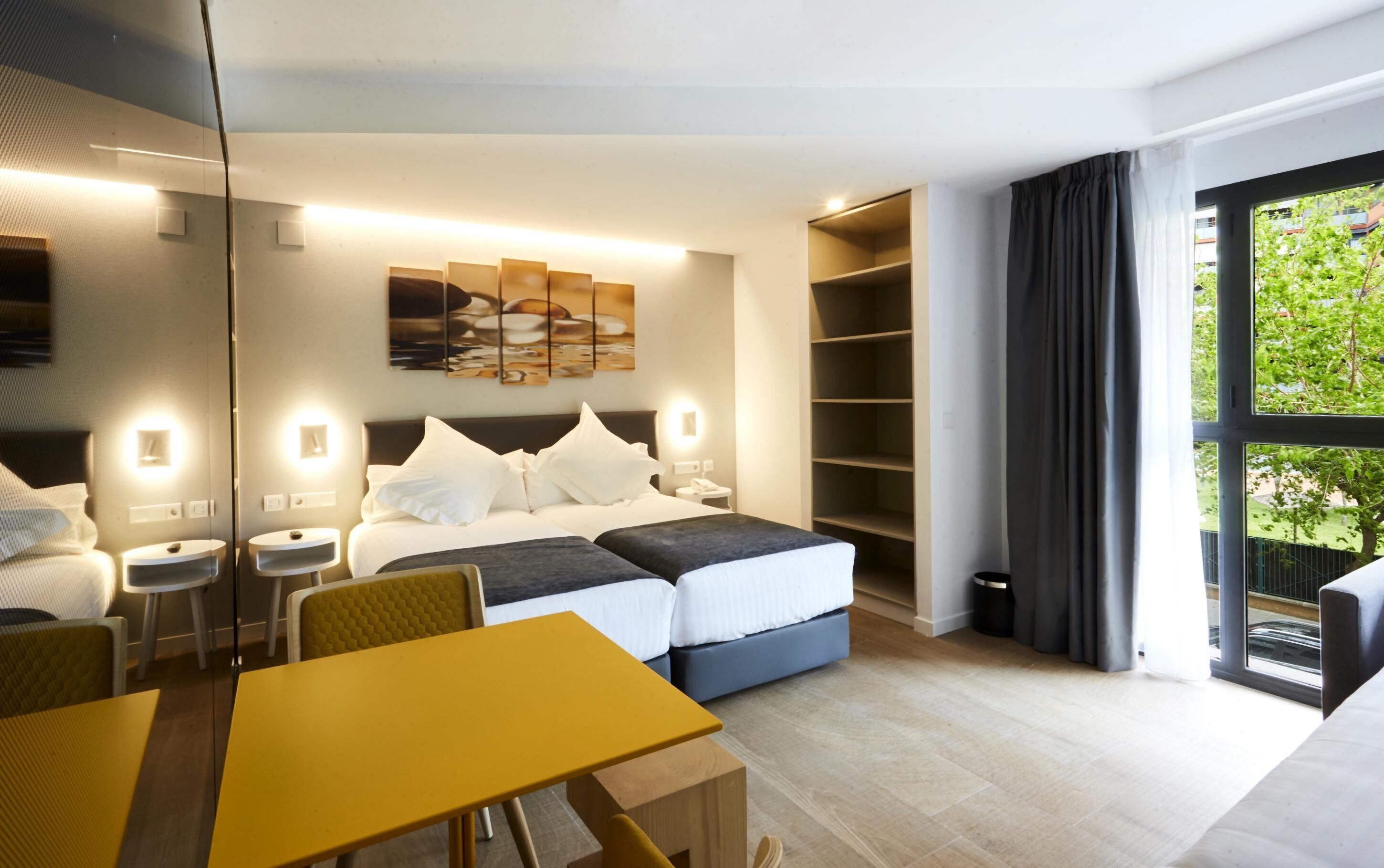 A modern hotel room with a bed, bedside tables, a desk, and a large window with curtains.