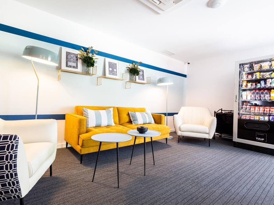 Un hall d’entrée moderne avec un canapé jaune, des fauteuils blancs, un distributeur automatique noir et des tableaux encadrés accrochés au mur.