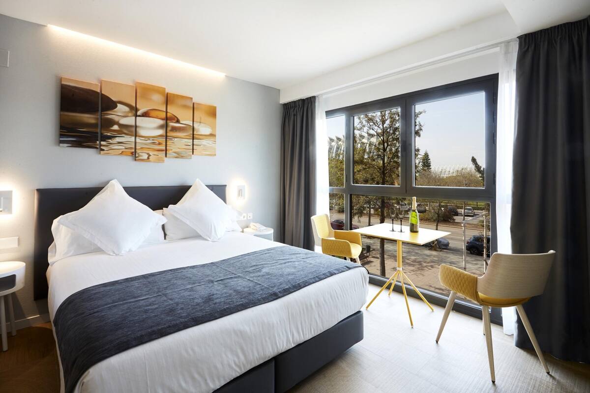 A modern hotel room with a large bed, a small table, two chairs, and a view of the outdoors.