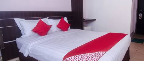 A hotel room with a bed, white bedding, a red and white patterned throw, a wooden headboard, a small bedside table, a framed picture on the wall, and a door with a handle.