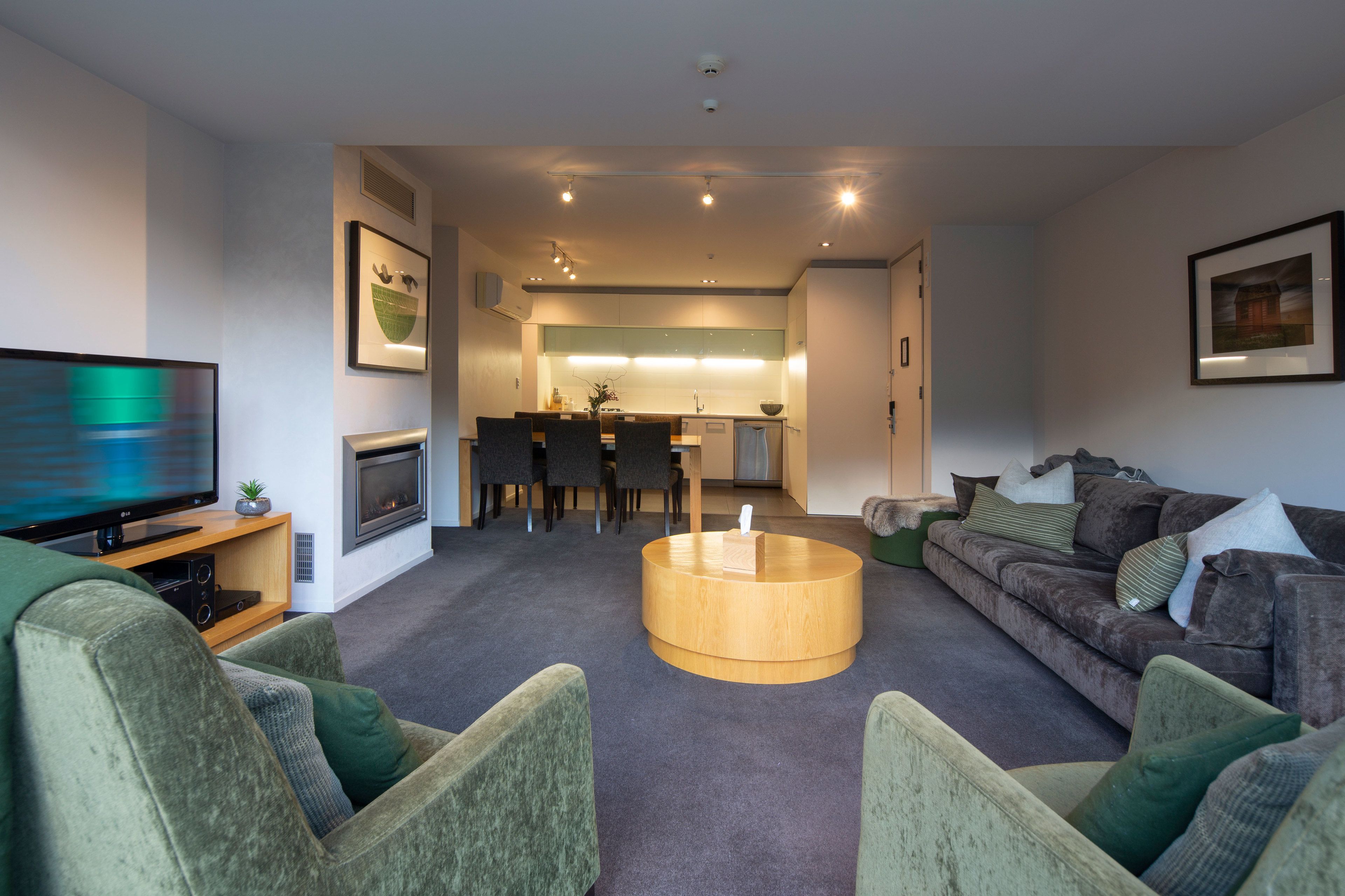 A modern living room with a flat-screen TV, green armchairs, a round coffee table, a sofa with cushions, and a kitchen area in the background.