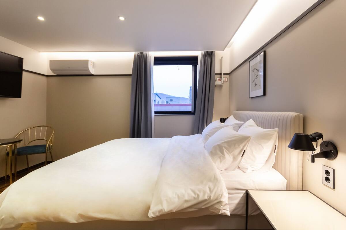 Habitación de hotel con cama, escritorio, silla, ventana con cortinas, unidad de aire acondicionado, lámpara de pared y un cuadro enmarcado.