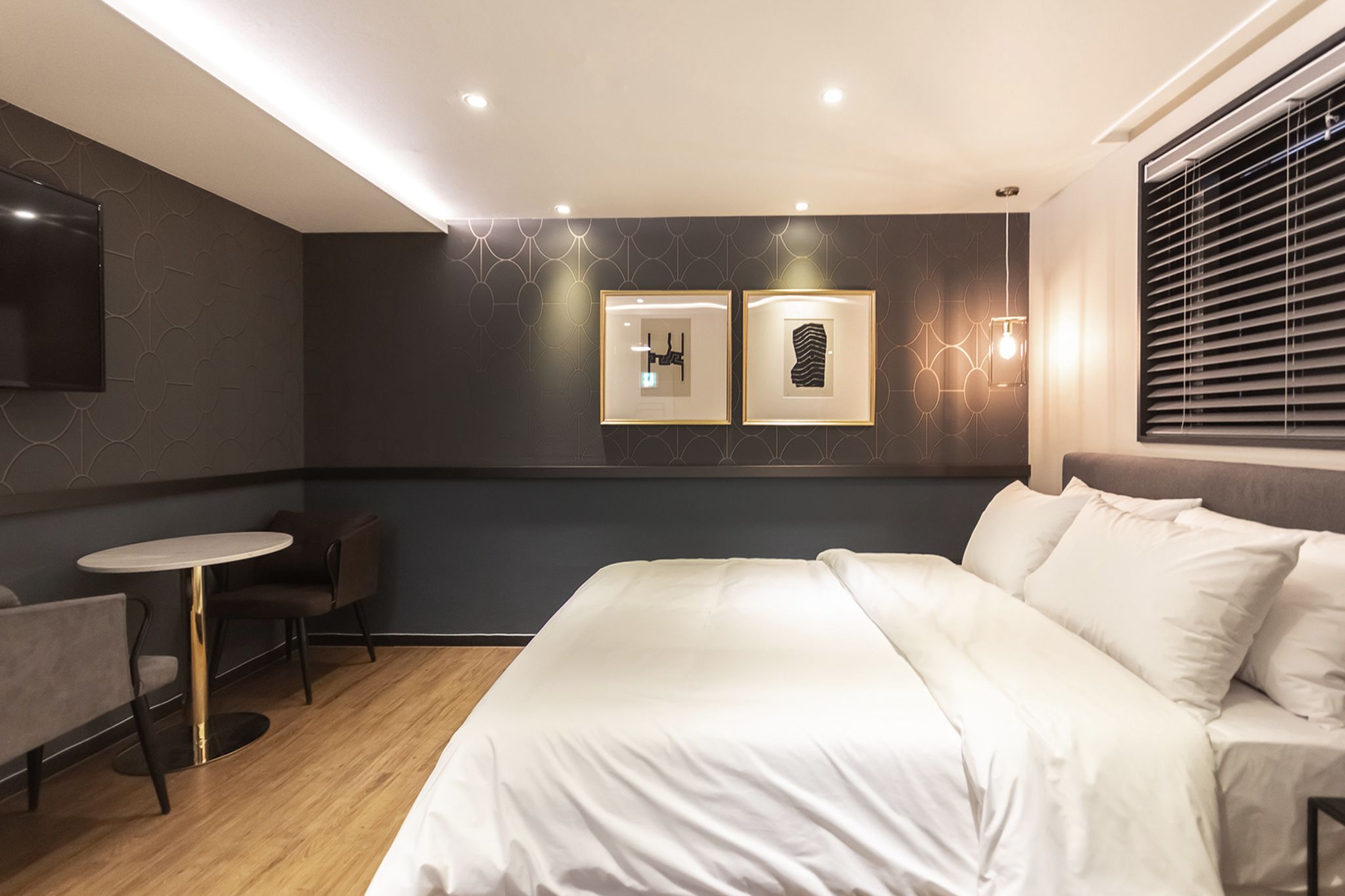 Moderni hotellihuone, jossa on suuri sänky, pieni pyöreä pöytä, kaksi tuolia, televisio ja kaksi kehystettyä kuvaa seinällä.