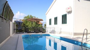 A swimming pool with lounge chairs and an umbrella, surrounded by a white building with green shutters.