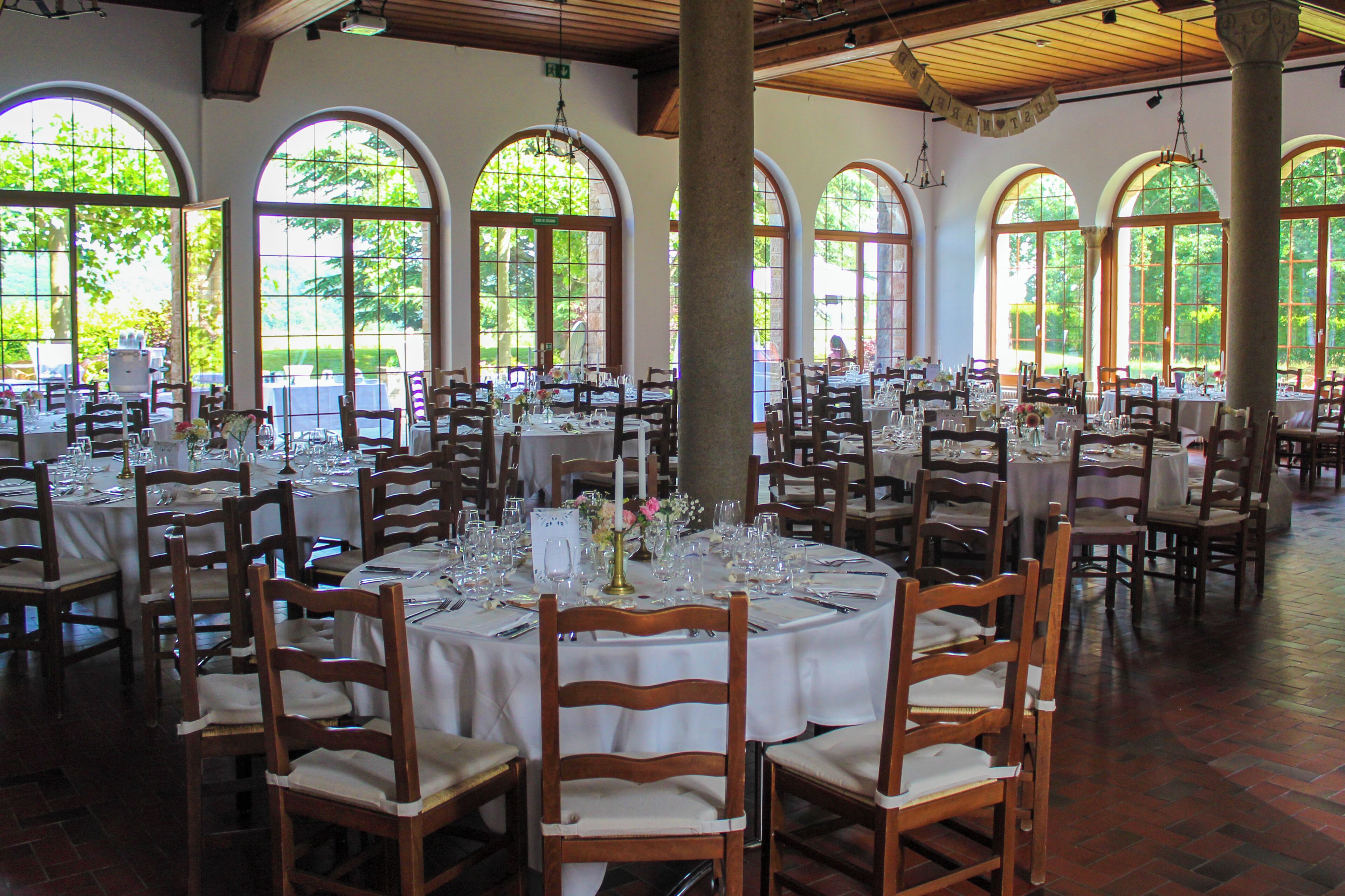 Un comedor amplio con varias mesas redondas dispuestas para eventos, ventanas arqueadas grandes y sillas de madera.