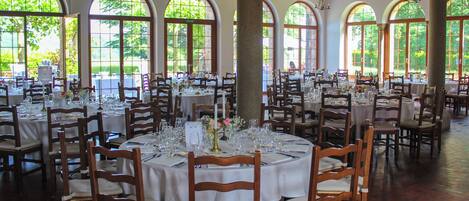Un comedor amplio con varias mesas redondas dispuestas para eventos, ventanas arqueadas grandes y sillas de madera.