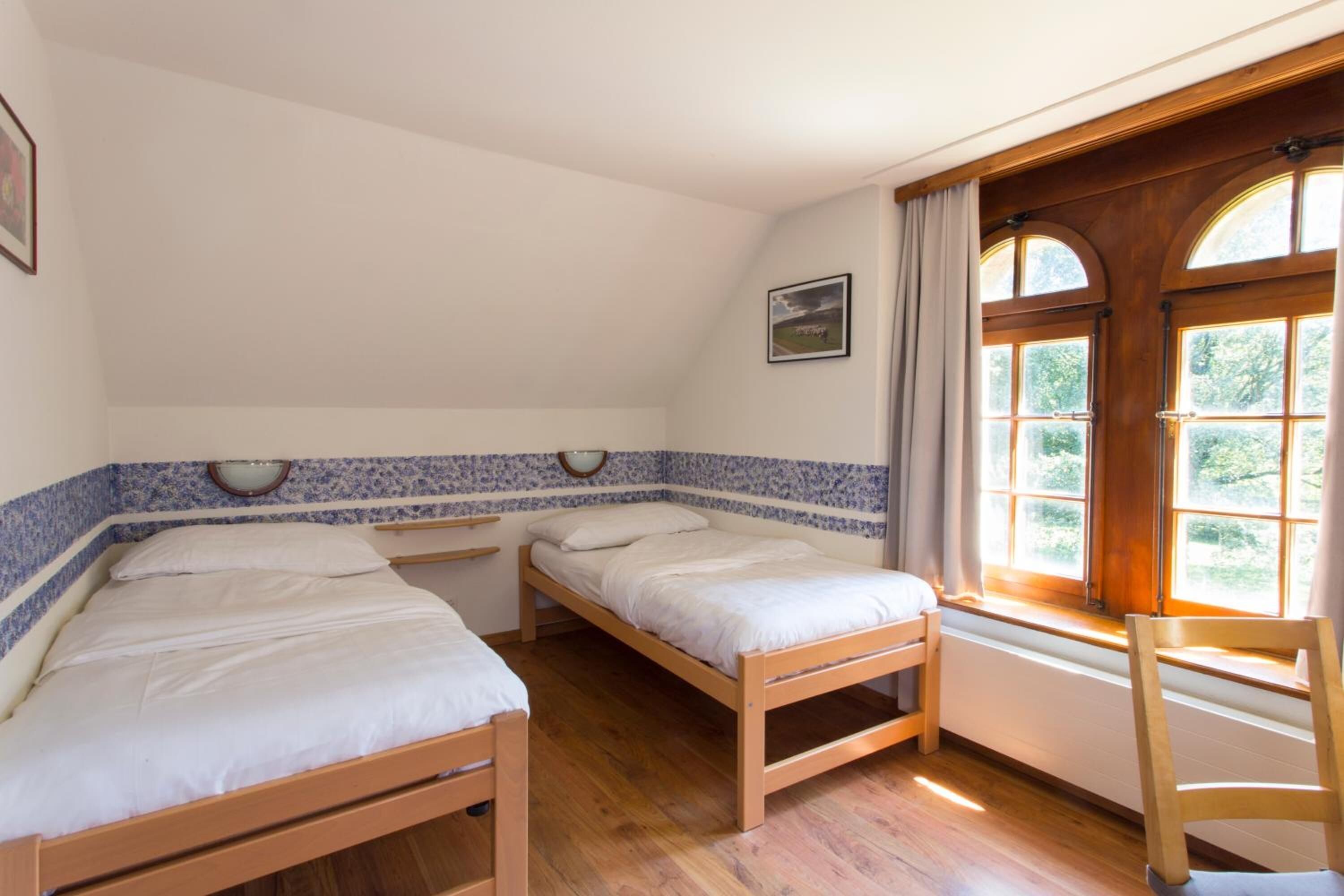 A room with two wooden beds, a window with curtains, and a wooden chair.