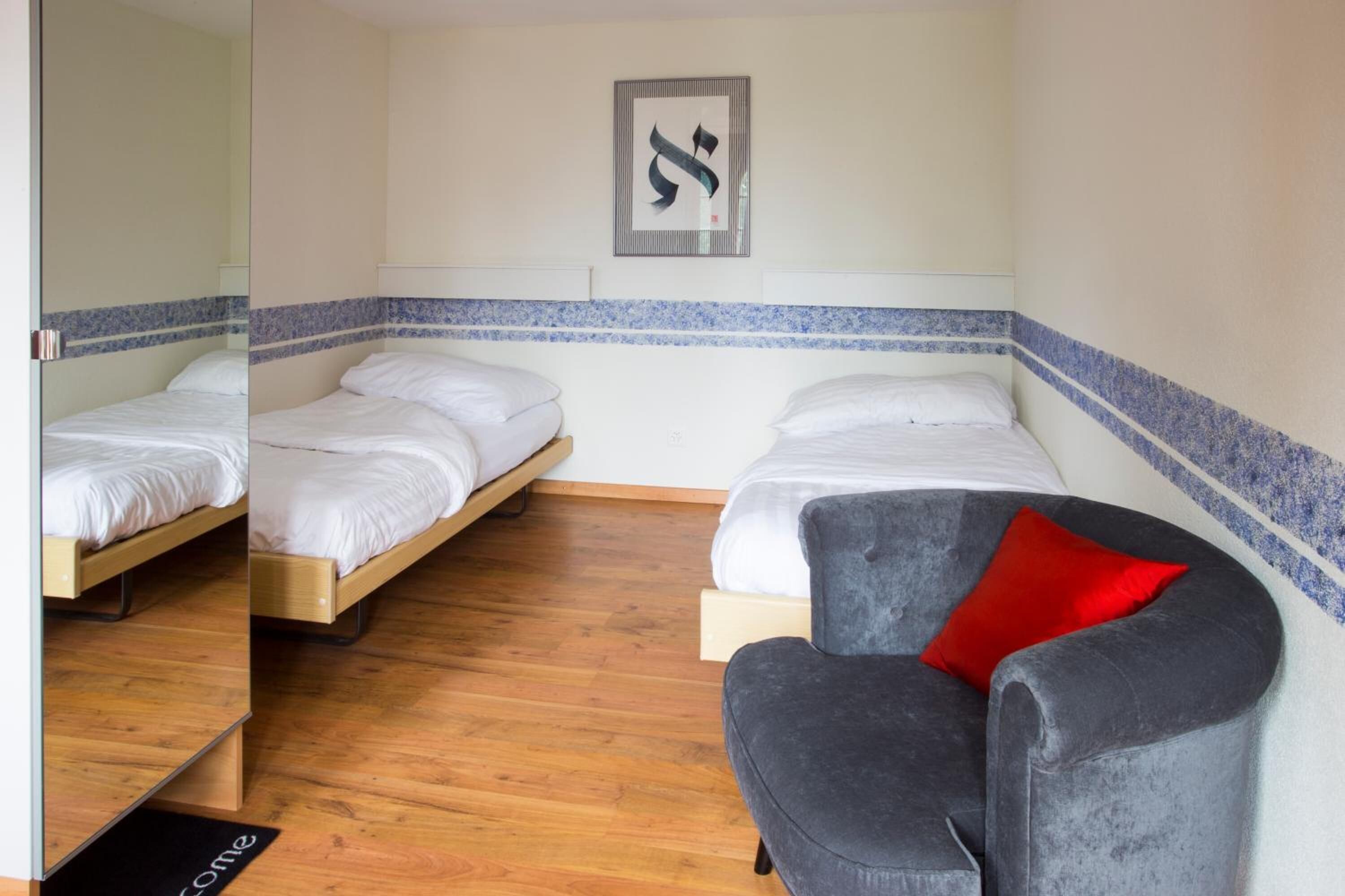 Une chambre avec deux lits simples, un fauteuil gris avec un coussin rouge et un paillasson.