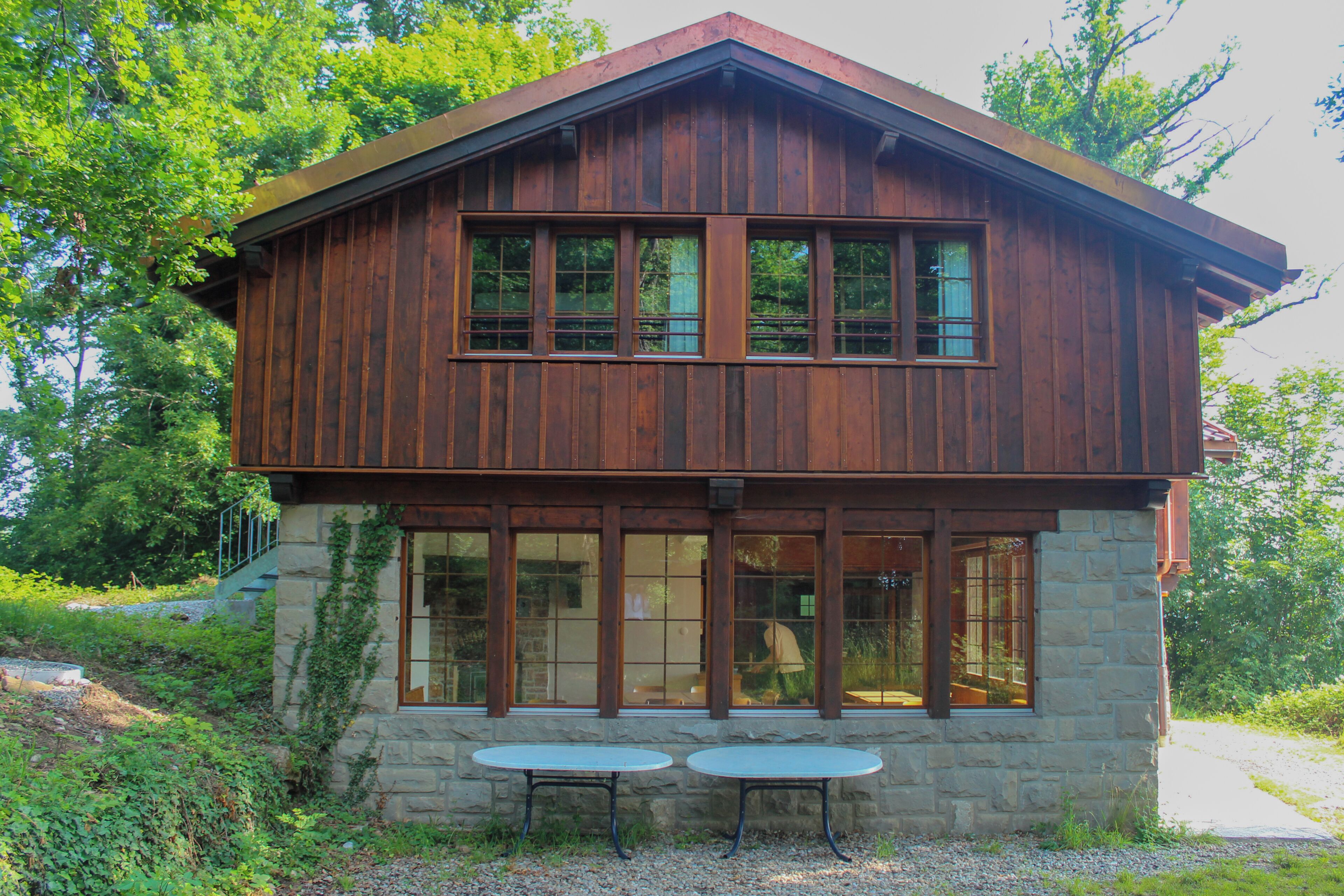 Eine zweistöckige Holzhütte mit Steinfundament, großen Fenstern und zwei Tischen im Freien.