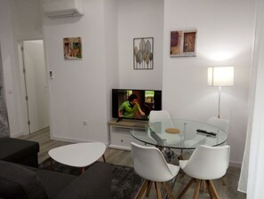 Moderni olohuone, jossa on lasinen ruokapöytä, valkoiset tuolit, litteänäyttöinen televisio ja sohva.