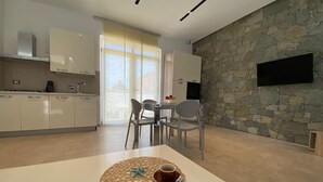 A modern kitchen with white cabinets, a dining area with a table and chairs, and a stone wall with a mounted TV. - VILLA BIANCA LOFT COMPLEX 1 (La Maddalena)